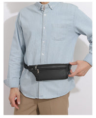 Black leather waist pack worn around the hips, front view with hand holding the bag to show shape